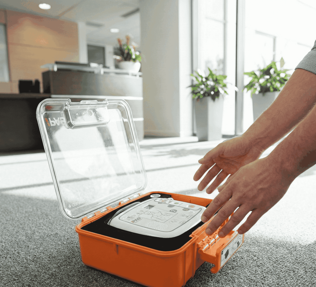 Ein geöffneter oranger Notfallkoffer mit einem iPAD CU-SP1 Defibrillator (AED) auf einem Büroteppich, während eine Person danach greift.
