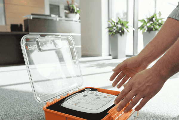 Ein geöffneter oranger Notfallkoffer mit einem iPAD CU-SP1 Defibrillator (AED) auf einem Büroteppich, während eine Person danach greift.